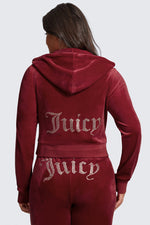 A woman in a plus-size OG Big Bling Velour Hoodie, maroon with thumbholes and rhinestone Juicy lettering on the back, stands facing away from the camera.