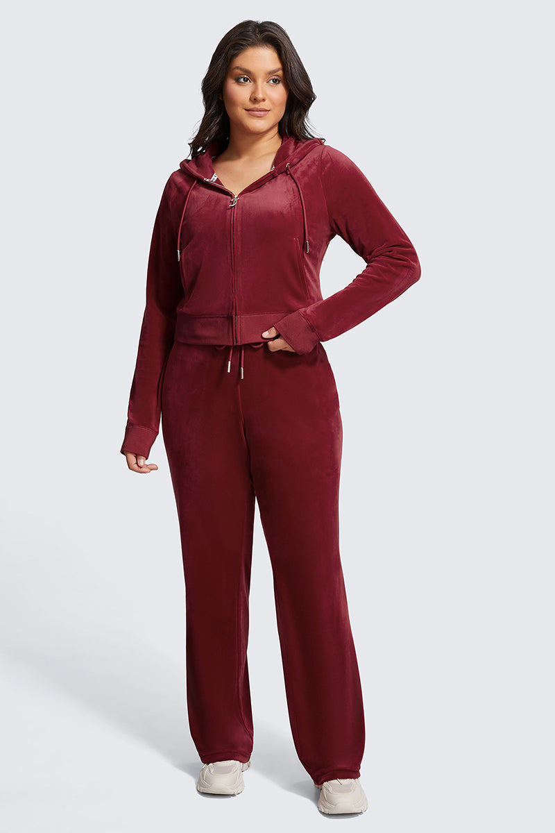 A woman with long dark hair wears the Plus-Size OG Big Bling Velour Hoodie in burgundy, featuring thumbholes and styled with matching wide-leg pants and white sneakers. She poses confidently against a plain background.