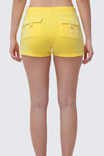Someone stands against a plain light gray background, seen from the waist down and behind, wearing Heritage Cotton Velour Embroidered Shorts in bright yellow with back pockets.