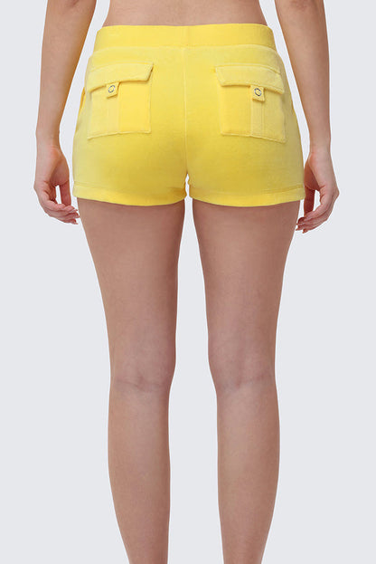 Someone stands against a plain light gray background, seen from the waist down and behind, wearing Heritage Cotton Velour Embroidered Shorts in bright yellow with back pockets.