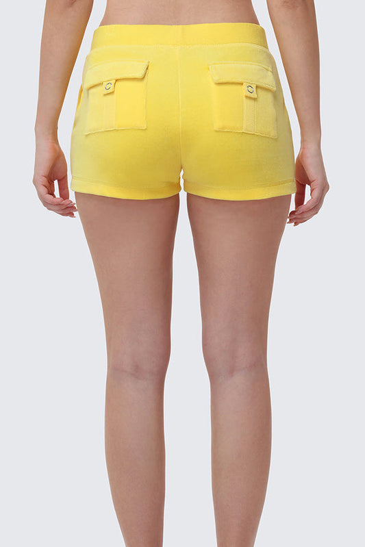 Someone stands against a plain light gray background, seen from the waist down and behind, wearing Heritage Cotton Velour Embroidered Shorts in bright yellow with back pockets.