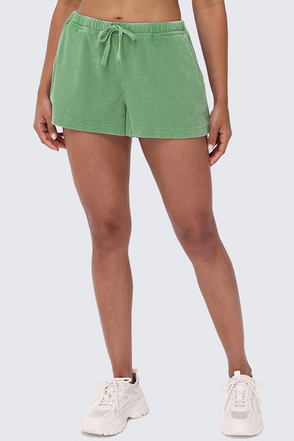 A person models the Big Bling Washed Terry Shorts in light green, featuring a rhinestone logo, paired with white sneakers. Only their lower torso and legs are shown against a plain, light background.