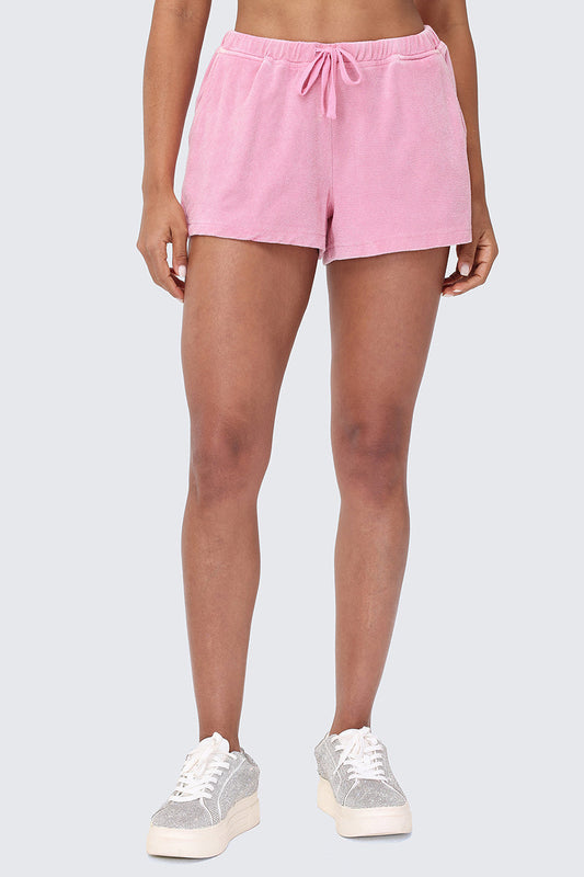 A person models the Big Bling Washed Terry Shorts in light pink with a drawstring waistband, paired with silver glitter sneakers, against a plain light gray background. The image is cropped at the midriff and below the knees.