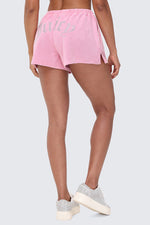 Facing away, a person wears Big Bling Washed Terry Shorts in pink with a rhinestone logo on the back and a comfy drawstring waistband. Glittery silver platform sneakers complete the look against a light gray background.