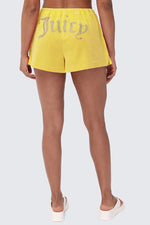 A person with brown skin faces away, wearing Big Bling Washed Terry Shorts in bright yellow featuring a rhinestone Juicy logo on the back, paired with white slide sandals against a light gray background.