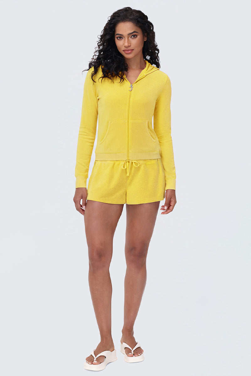 A woman with curly hair and minimal makeup faces forward, wearing Big Bling Washed Terry Shorts in bright yellow paired with a matching zip-up hoodie and white sandals against a plain light background.