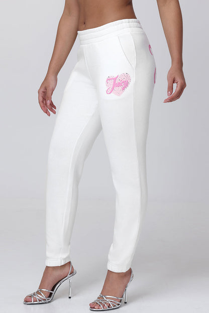 A person models Sweetheart Fleece Sweatpants featuring a pink graphic, paired with silver high-heeled sandals, standing against a plain light gray background. The relaxed fit reveals their midriff and arms.