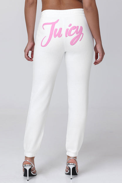 A person stands facing away, wearing Sweetheart Fleece Sweatpants in a relaxed fit with "Juicy" in pink script across the back, paired with white high-heeled sandals and a bare back.