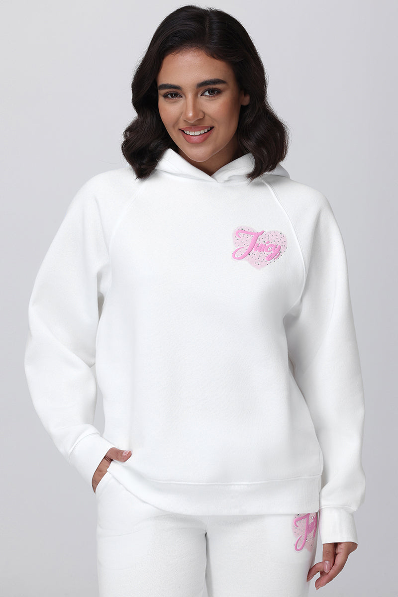 A woman with dark hair smiles in the Sweetheart Fleece Oversized Hoodie and matching white sweatpants featuring a pink, sparkling J logo, posing against a plain light background for a cozy, casual look.