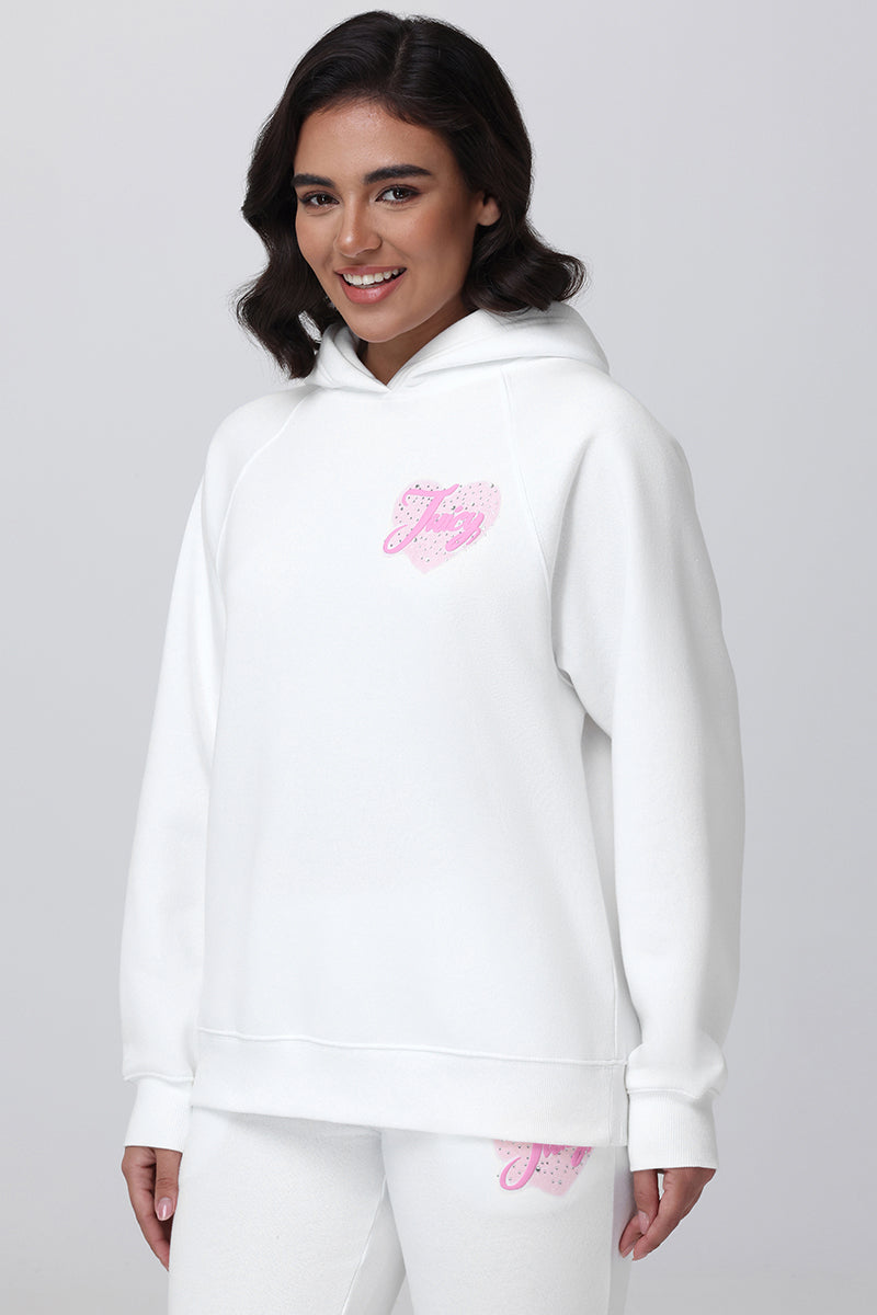 A woman with shoulder-length dark hair smiles while wearing the Sweetheart Fleece Oversized Hoodie and matching sweatpants, both white with a small pink graphic, standing out against a plain light gray background.