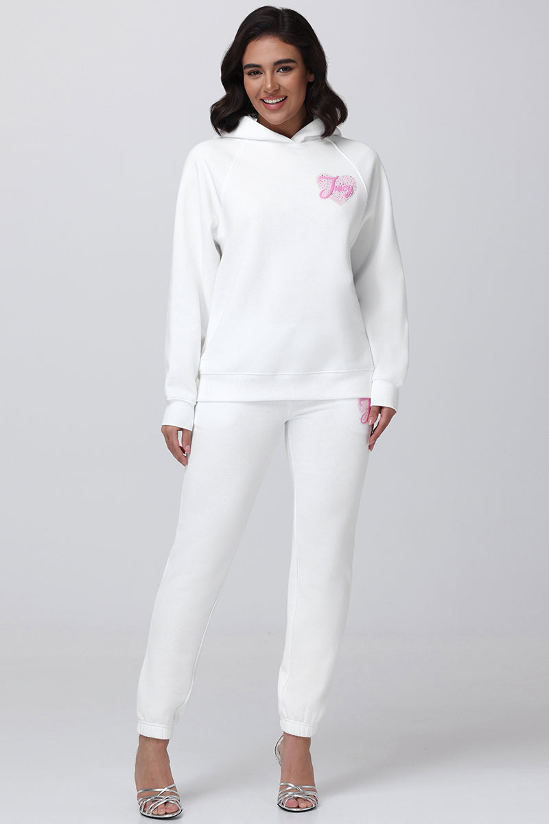 A woman stands smiling in the Sweetheart Fleece Oversized Hoodie and matching jogger pants, both white with pink embroidery, styled with silver high-heeled sandals for a chic casual look against a light gray background.