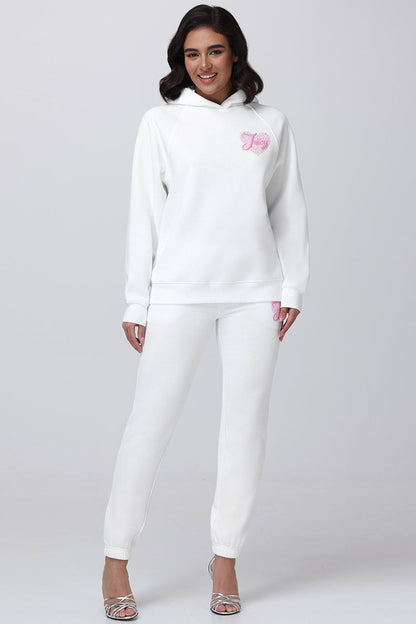 A woman stands smiling in the Sweetheart Fleece Oversized Hoodie and matching jogger pants, both white with pink embroidery, styled with silver high-heeled sandals for a chic casual look against a light gray background.