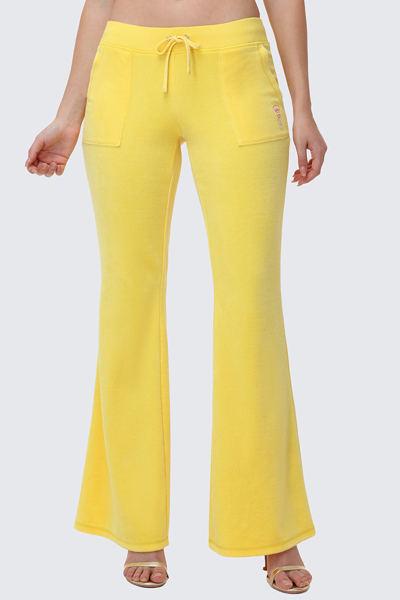 A person in bright yellow Heritage Cotton Velour Track Pants with front pockets and a drawstring waist stands against a plain light gray background, wearing open-toe heels.