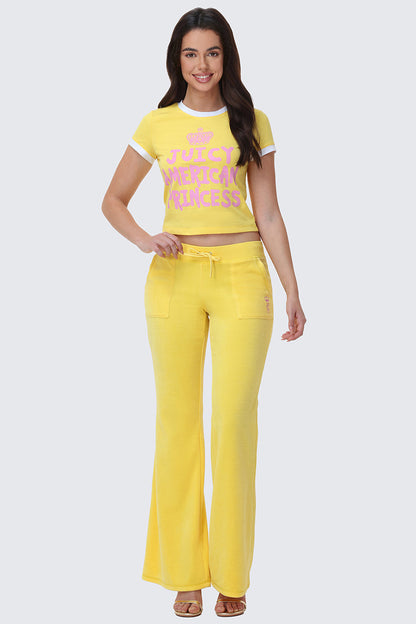 A woman smiles with her hands by her sides, wearing yellow Heritage Cotton Velour Track Pants and a matching t-shirt, standing against a plain light background.