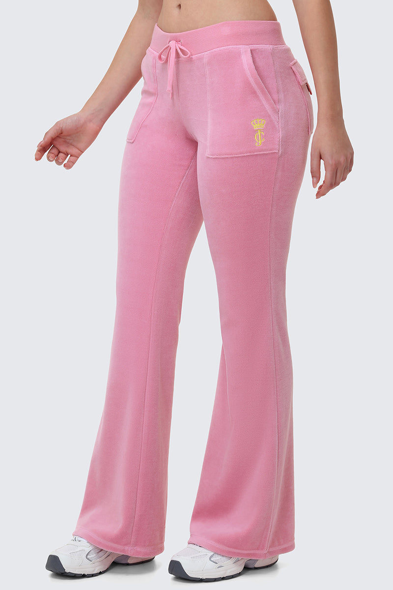 A person wearing Heritage Cotton Velour Track Pants in light pink with a drawstring waist, front pockets, and a small yellow crown logo, paired with white sneakers, shown from the waist down.