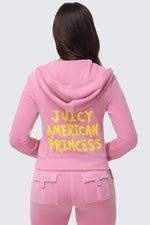 A woman with long brown hair is seen from behind wearing the American Princess Heritage Hoodie, a pink velour zip-up with "JUICY AMERICAN PRINCESS" printed in bold yellow letters on the back.