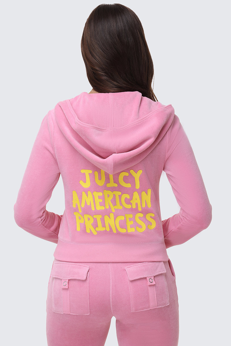 A woman with long brown hair is seen from behind wearing the American Princess Heritage Hoodie, a pink velour zip-up with "JUICY AMERICAN PRINCESS" printed in bold yellow letters on the back.