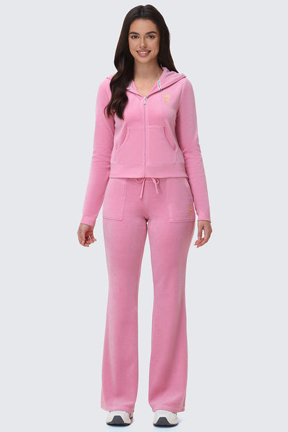 A woman faces forward, smiling, wearing the American Princess Heritage Hoodie in pink velour with a crown logo, matching pink flared pants, and white sneakers against a plain light gray background.