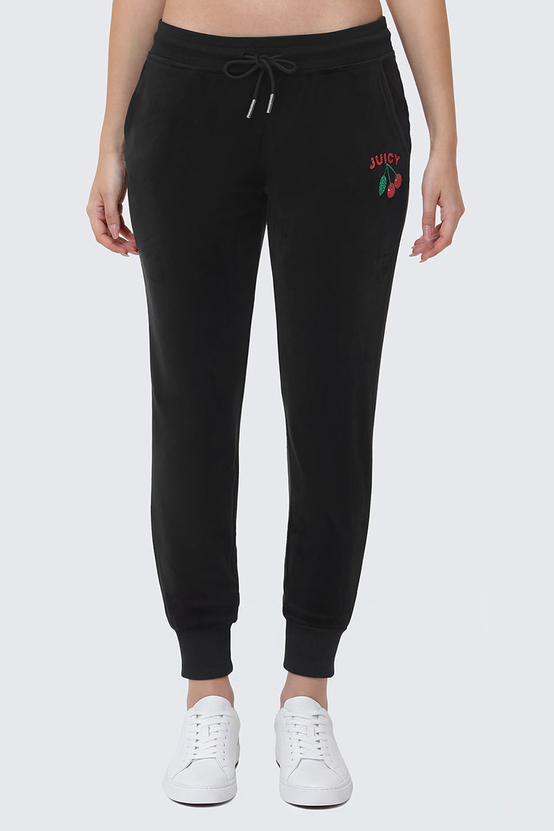 Someone wears the Cherry Bling Velour Jogger—black velour joggers with a drawstring waist, red JUICY embroidery, and a cherry logo on the left hip—paired with white sneakers.