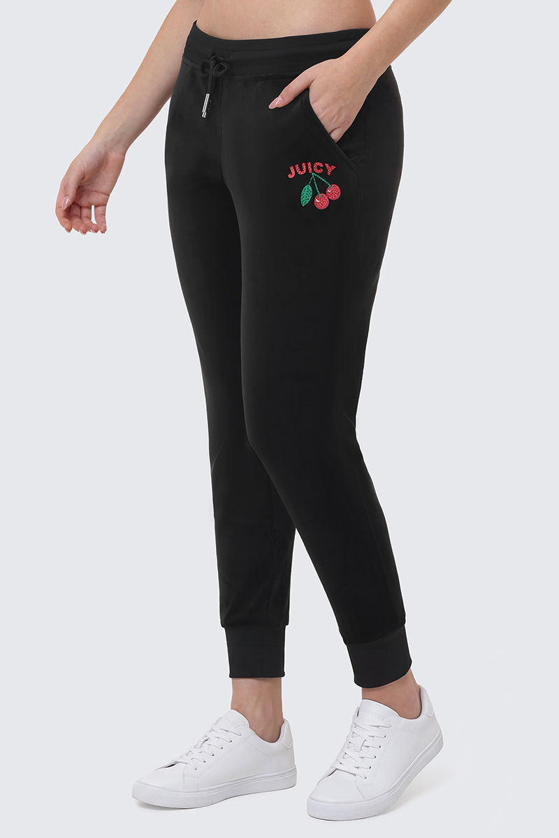 Someone wearing the Cherry Bling Velour Jogger, featuring a drawstring waist, side pockets, and a Juicy cherry logo on the left thigh, styled with white sneakers.