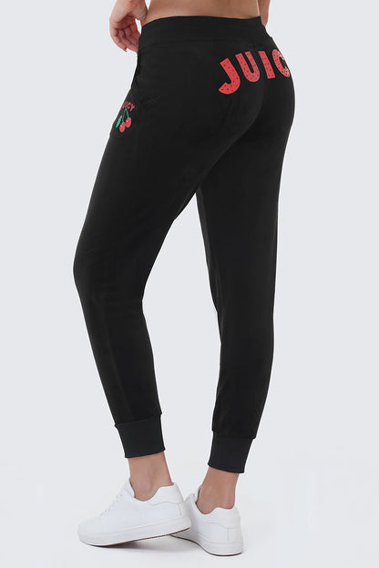 Wearing Cherry Bling Velour Joggers with a pink Juicy graphic on the back and a cherry logo on the side, paired with white sneakers, the person stands against a light background.