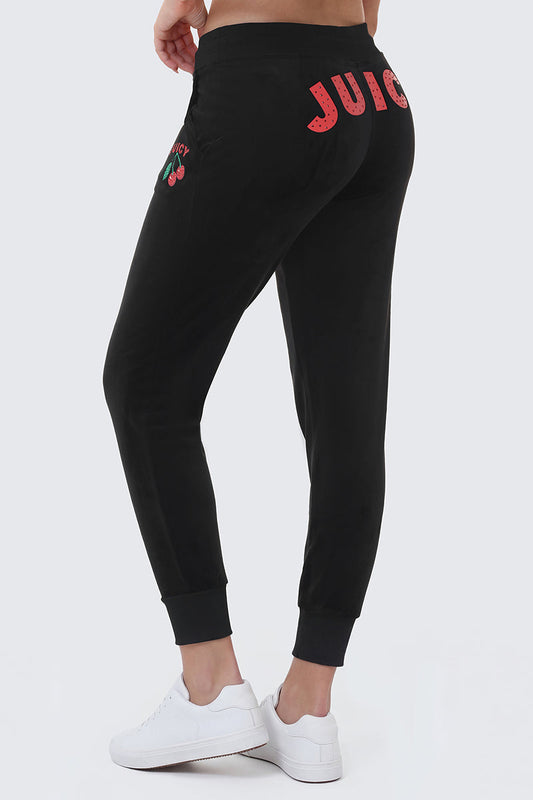 Wearing Cherry Bling Velour Joggers with a pink Juicy graphic on the back and a cherry logo on the side, paired with white sneakers, the person stands against a light background.