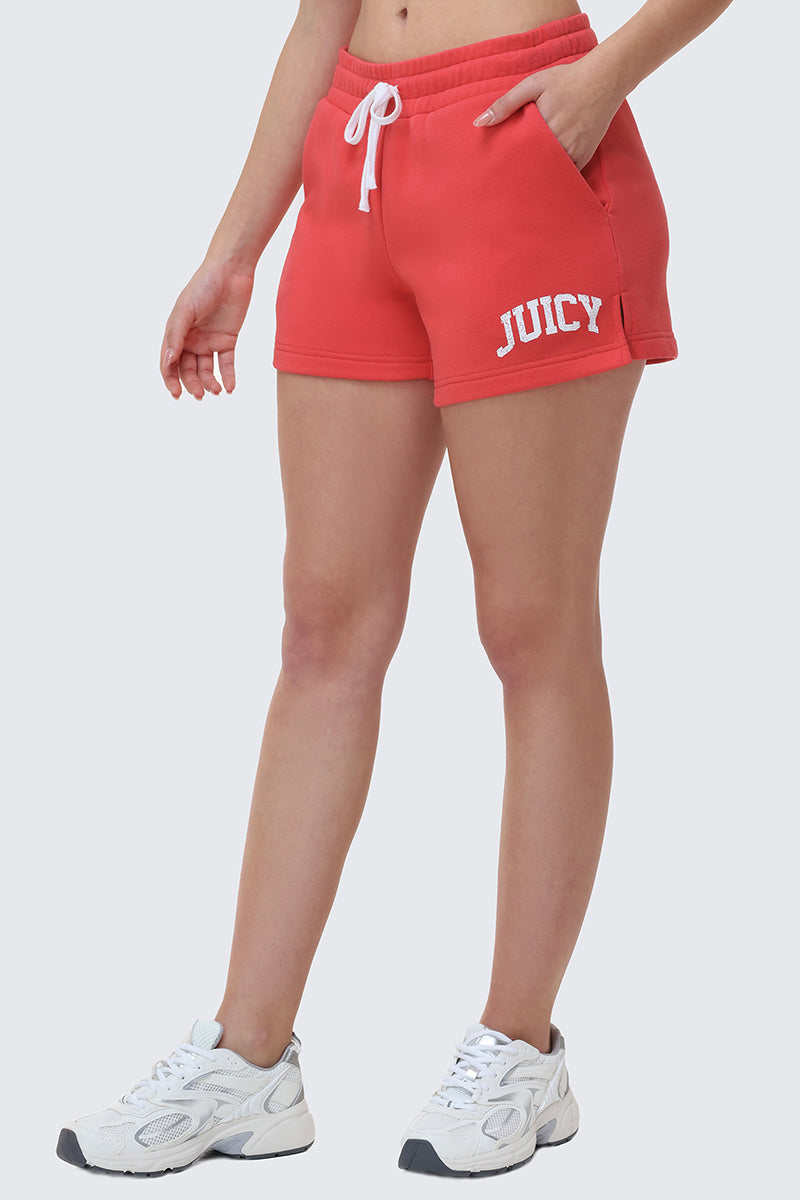Someone stands against a plain light background, wearing bright red Collegiate Booty Shorts with the Juicy logo on the left leg, a white drawstring waist, and white athletic shoes.