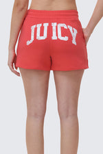 A person is shown from the waist down, wearing Collegiate Booty Shorts in red terry with a drawstring waist and "JUICY" printed in large white letters across the back.