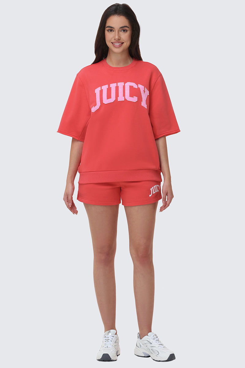 A woman smiles, modeling Collegiate Booty Shorts and a loose red Juicy logo T-shirt with white sneakers, standing against a plain light gray background.