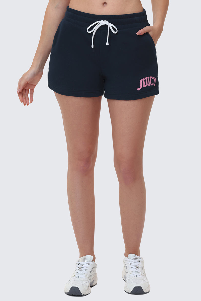 A person wears Collegiate Booty Shorts in navy blue with a white drawstring waist and pink JUICY logo on the left, paired with white sneakers. The upper body is not visible.