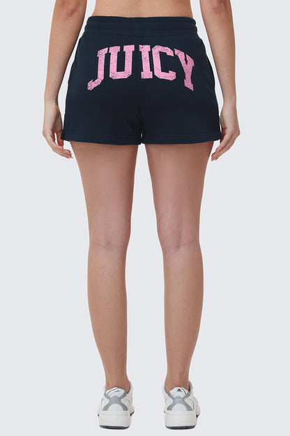 A person is seen from behind wearing Collegiate Booty Shorts in black terry with a bold pink Juicy logo across the back, paired with white sneakers.