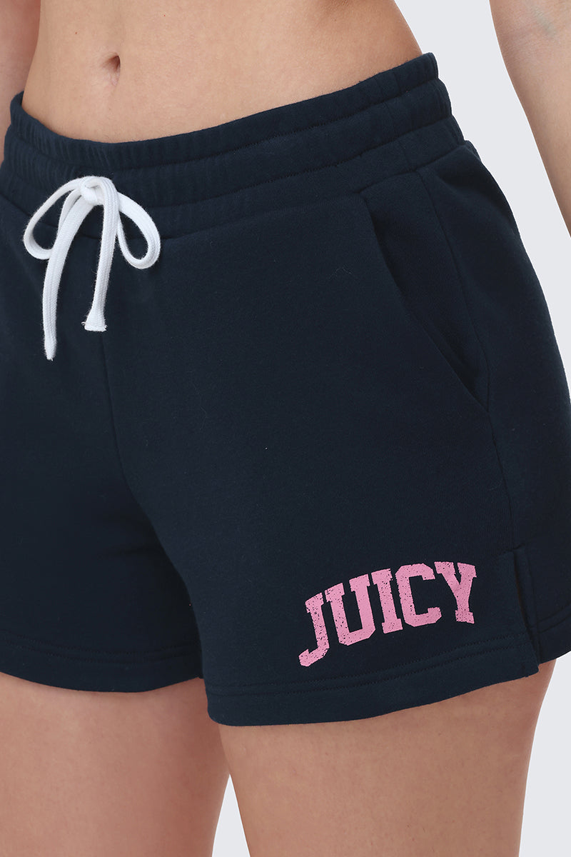 Close-up of a person wearing Collegiate Booty Shorts in black terry, featuring a white drawstring waist and pink Juicy logo on the left thigh. The image shows their torso and part of their legs.