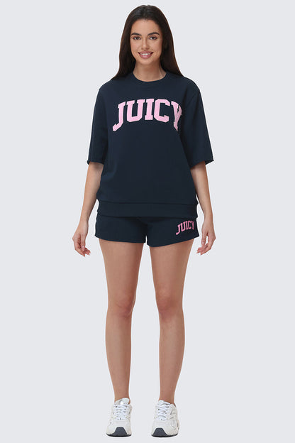 A woman smiles facing forward, wearing navy Collegiate Booty Shorts with a drawstring waist and pink Juicy logo, a matching top with a large pink Juicy logo, and white sneakers.