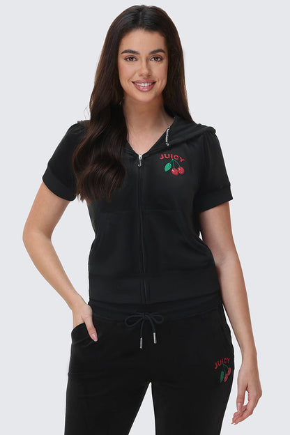 A woman with long brown hair smiles, wearing the Cherry Bling Velour Hoodie and matching pants with red Juicy cherry graphics, standing with one hand in her pocket against a light background.