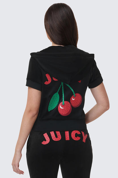 A woman faces away from the camera in the Cherry Bling Velour Hoodie, showcasing a juicy cherry graphic and JUICY on the back, paired with black pants featuring rhinestone-studded JUICY lettering across the seat.