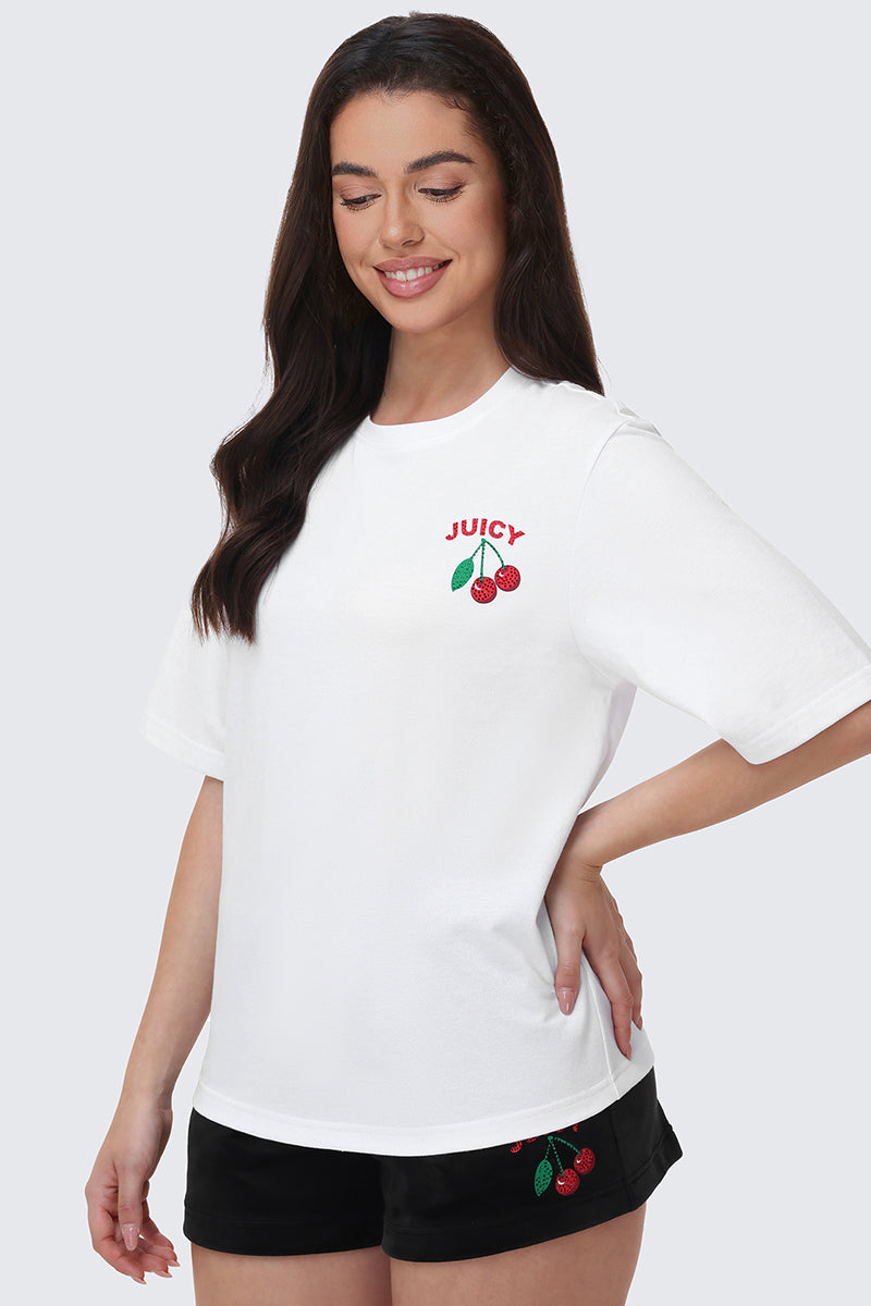 A smiling woman with long dark hair wears the Cherry Bling Graphic Tee—featuring a bold JUICY cherry graphic with rhinestone accents—paired with black shorts with a small cherry motif. She stands against a light background, hand on hip.
