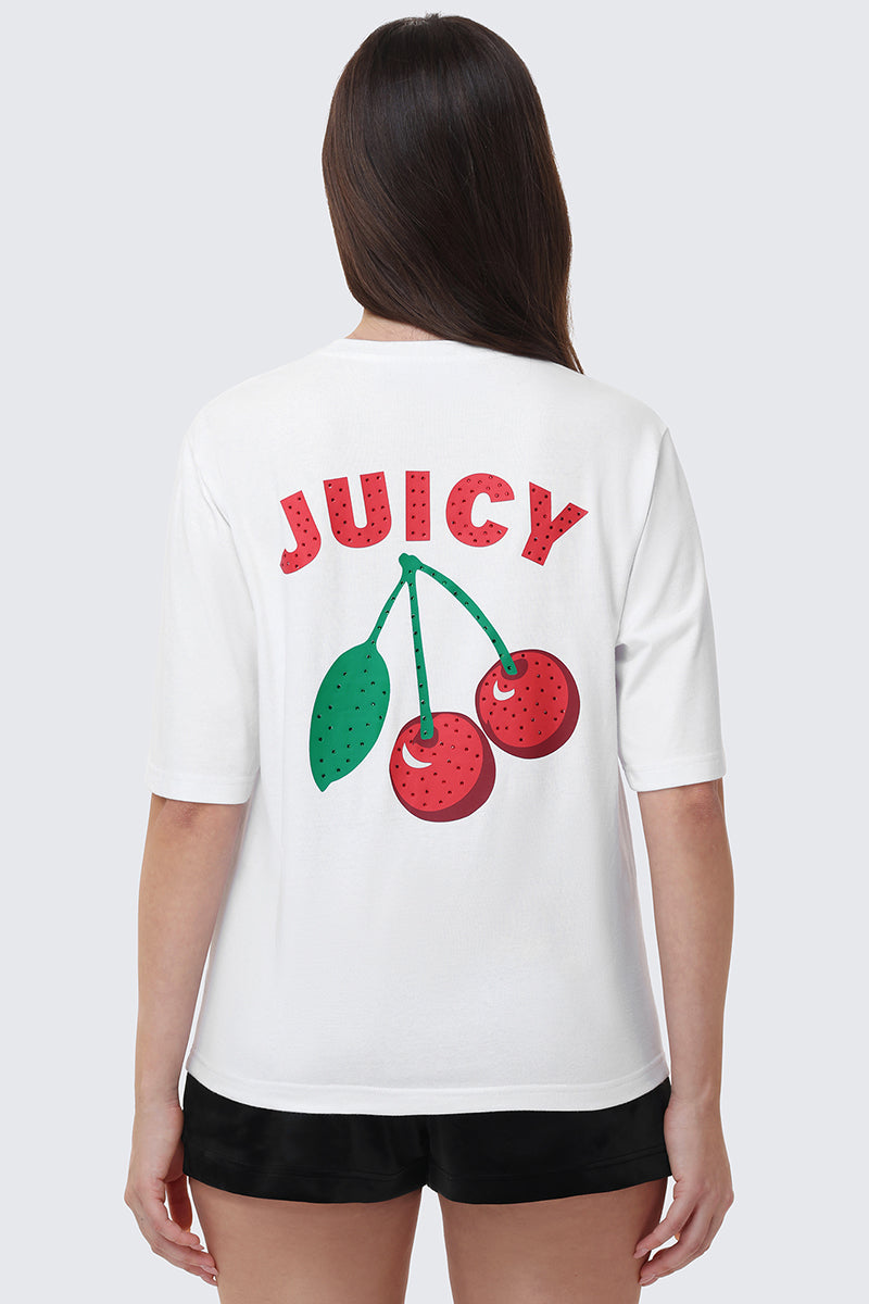 A person with long brown hair wears the Cherry Bling Graphic Tee—a white relaxed fit with a bold JUICY print and two large red cherries with a green leaf on the back—styled with black shorts.