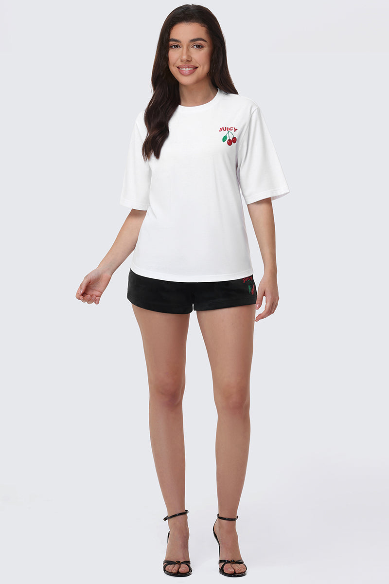 A woman smiles in the Cherry Bling Graphic Tee featuring a rhinestone cherry graphic on the left chest, paired with black shorts and strappy black heels, against a plain light gray background.