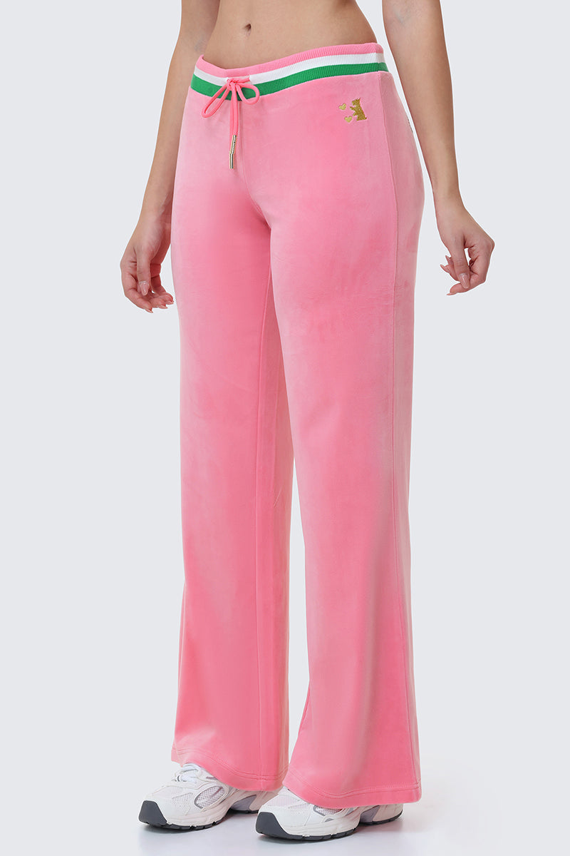 Wearing Heritage Stripe Bling Velour Track Pants in pink with a green and white striped waistband, drawstring, and white sneakers, the look spotlights casual style and subtly showcases the Juicy brand on the lower half.