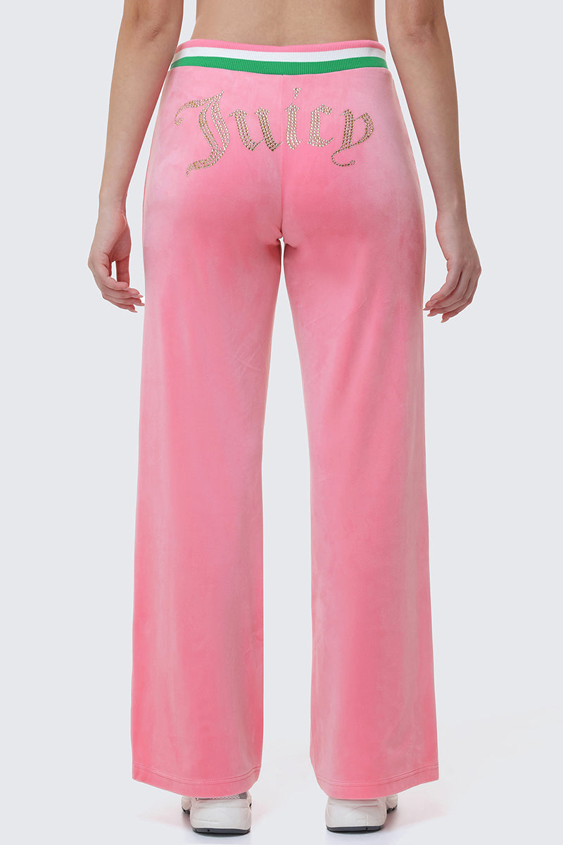 Wearing Heritage Stripe Bling Velour Track Pants, which feature pink velour, a Juicy rhinestone logo on the back, and a green and white striped waistband, the person stands against a light gray background.