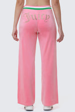 Wearing Heritage Stripe Bling Velour Track Pants, which feature pink velour, a Juicy rhinestone logo on the back, and a green and white striped waistband, the person stands against a light gray background.