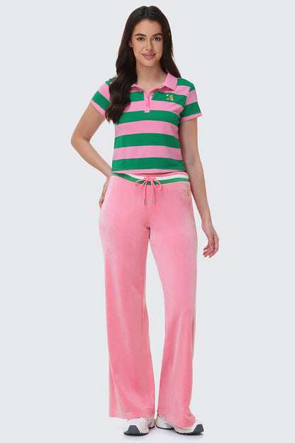 A woman smiles in Heritage Stripe Bling Velour Track Pants, a green and pink striped polo shirt, and white sneakers against a plain light background.