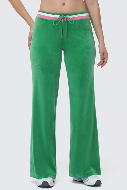A person is wearing Heritage Stripe Bling Velour Track Pants, featuring a wide-leg green velour design with a striped sporty waistband and drawstring, paired with white sneakers. Only the lower half of the body is visible.