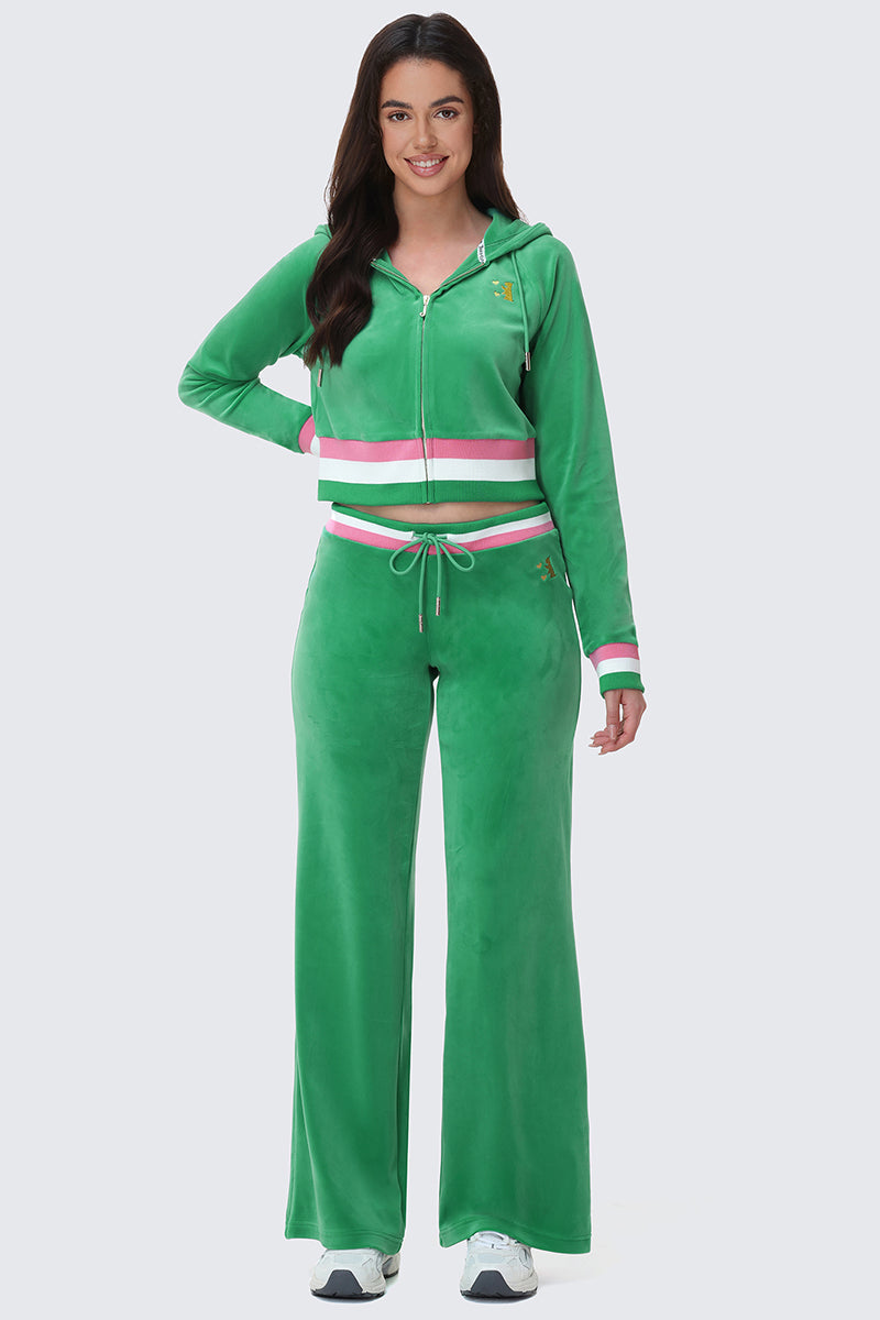 A woman smiles while wearing Heritage Stripe Bling Velour Track Pants with a sporty striped waistband and matching drawstring, standing forward with one hand on her hip to highlight the stylish details of her outfit.