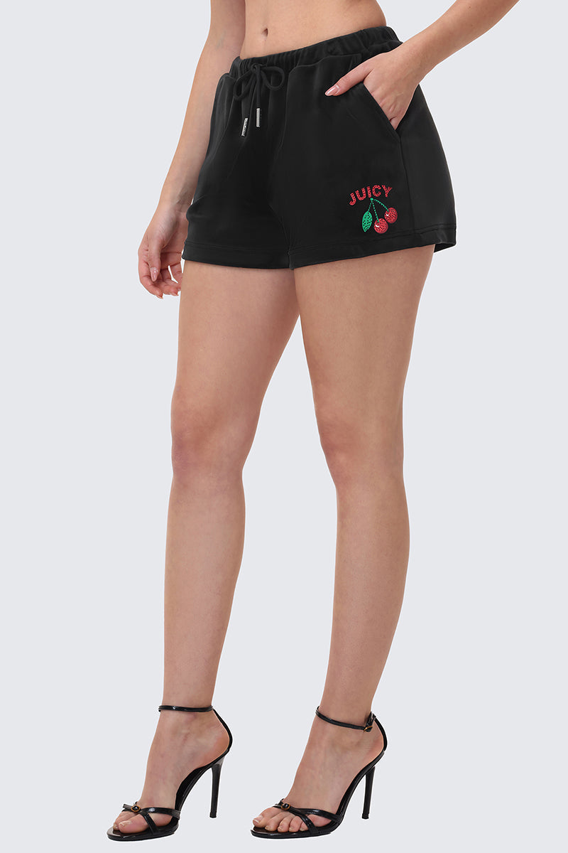 A person models the Cherry Bling Velour Shorts, featuring a Juicy graphic and cherry logo embroidery on the left leg, styled with black high-heeled sandals. Only the lower half of the body is shown against a plain background.