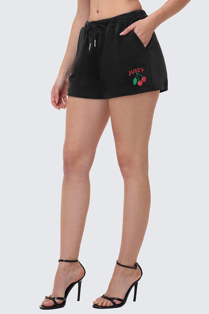 A person models the Cherry Bling Velour Shorts, featuring a Juicy graphic and cherry logo embroidery on the left leg, styled with black high-heeled sandals. Only the lower half of the body is shown against a plain background.