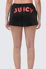 A person is seen from behind wearing Cherry Bling Velour Shorts featuring a bold Juicy graphic in large red letters across the back, set against a plain, light-colored background.