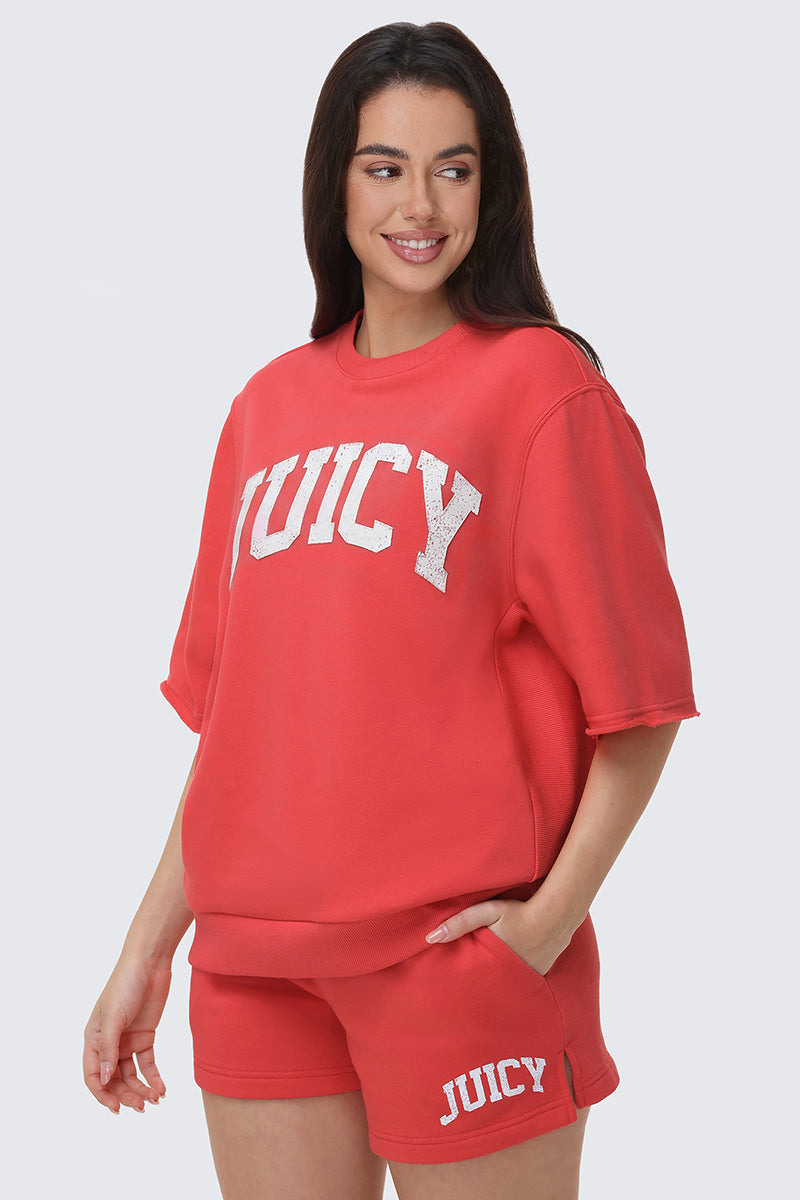 A woman with long dark hair smiles while wearing a red Collegiate Sweatshirt with “JUICY” in white letters and matching shorts, her hand in her pocket highlighting the relaxed fit of the set.
