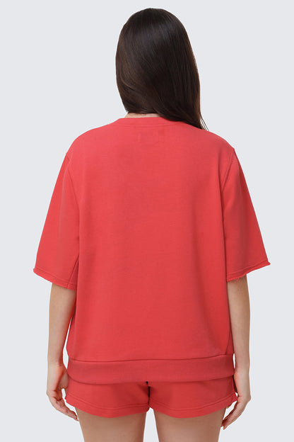 A person with long dark hair stands with their back to the camera, wearing a relaxed fit red Collegiate Sweatshirt and matching shorts, evoking a collegiate-style look against a plain light background.
