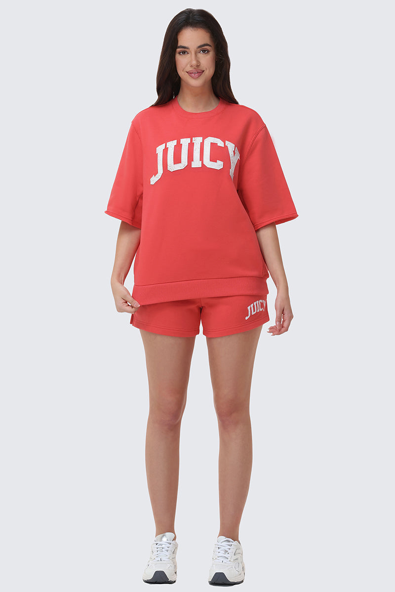 A woman faces forward, smiling, in a relaxed fit red Collegiate Sweatshirt with matching shorts and white sneakers against a plain light background.